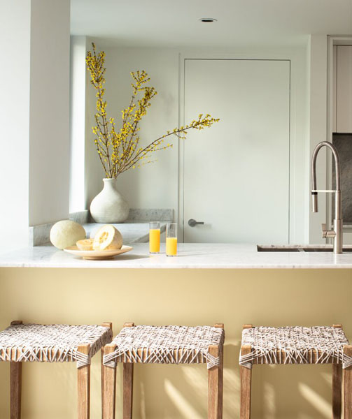 A kitchen with breakfast bar painted in Golden Straw 2152-70. The walls, woodwork and ceiling are painted in White Heron OC-57 and the room is styled with yellow flowers.